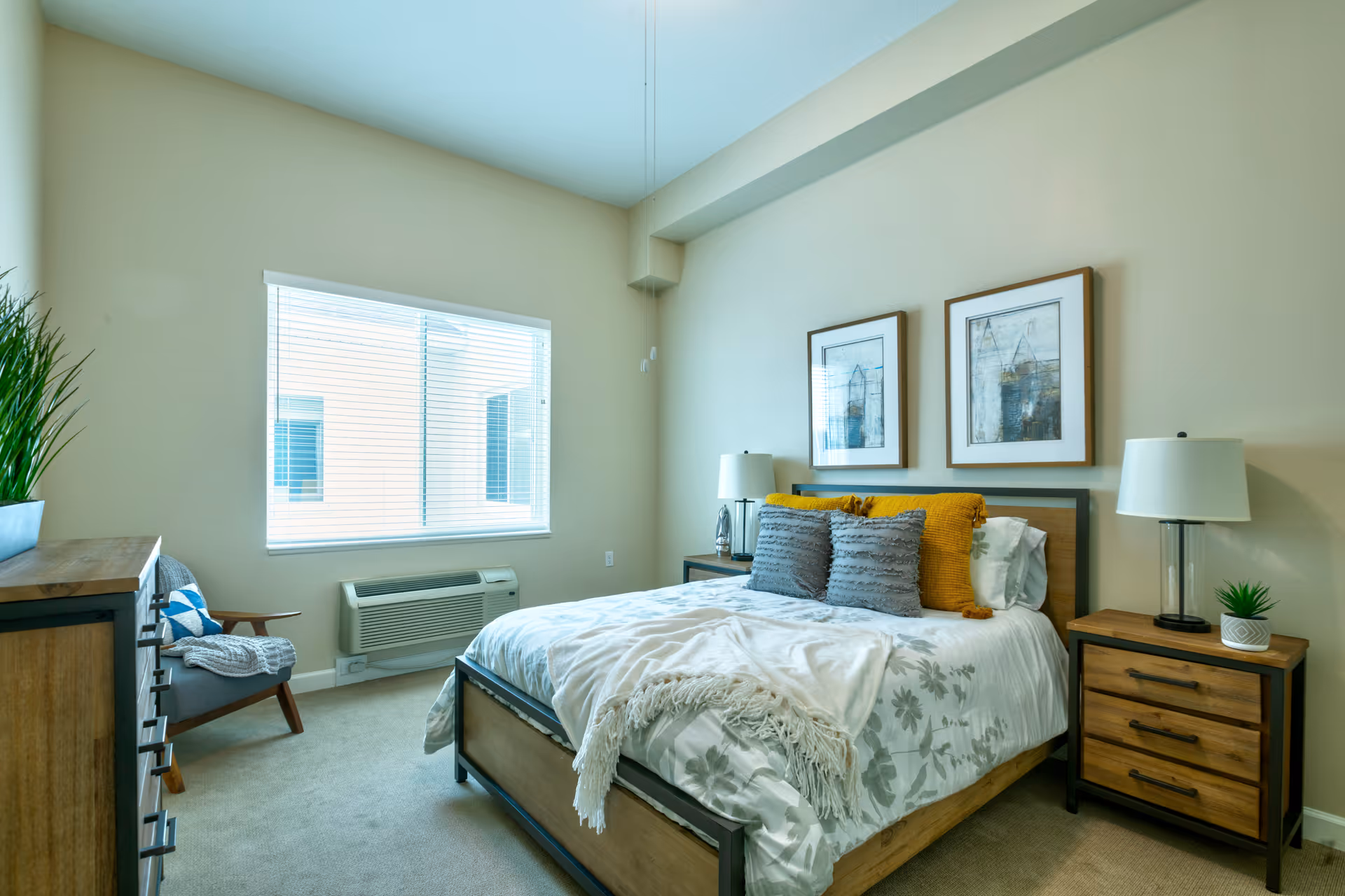 A bright, furnished bedroom with a bed, nightstands, dresser, armchair, and a window with blinds.