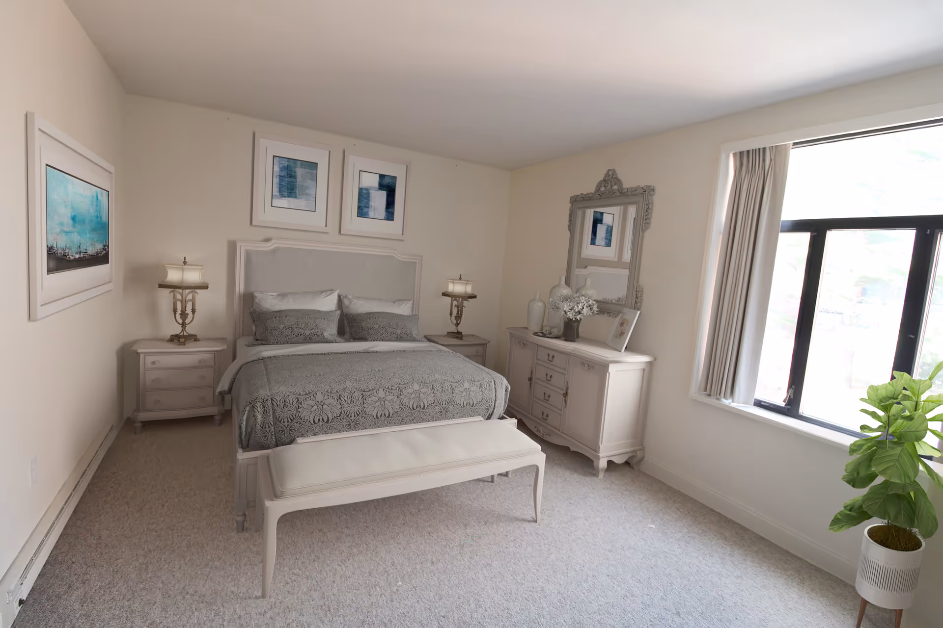 A bright bedroom with a large window on the right side letting in natural light. The room features a bed with a gray patterned bedspread and matching pillows, flanked by two nightstands each with a decorative lamp. Above the bed are two framed abstract artworks. On the right side of the room is a dresser with a large ornate mirror, decorative vases, and a framed photo. A cushioned bench sits at the foot of the bed, and a potted plant is placed near the window.