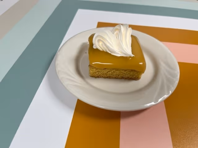A small square piece of cake with caramel-colored frosting topped with a swirl of white whipped cream on a white plate, placed on a surface with a colorful geometric pattern.