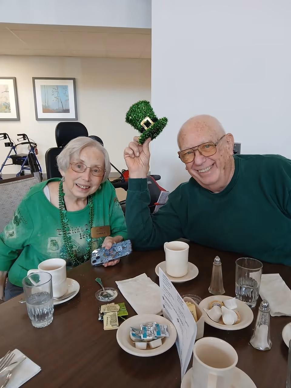 Two elderly people sitting at a table in a dining area. The woman on the left is wearing glasses, a green shirt, and green beaded necklaces, holding a phone. The man on the right is smiling, wearing glasses and a green sweater, holding a small green leprechaun hat above his head. On the table are cups, glasses of water, sugar packets, creamers, salt and pepper shakers, and napkins. In the background, there are framed pictures on the wall and a walker.