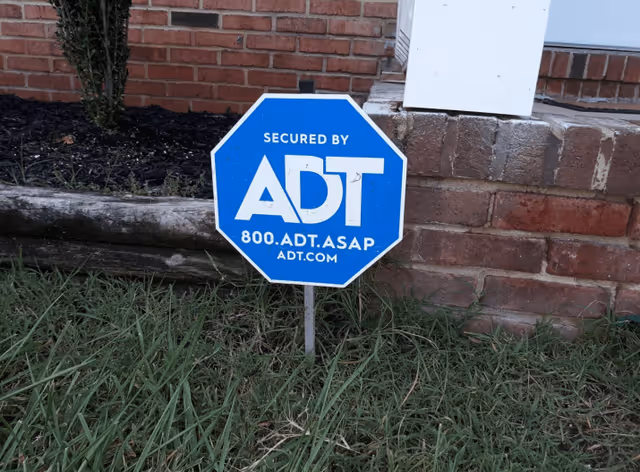 Blue octagonal ADT security sign staked in grass in front of a brick building foundation.
