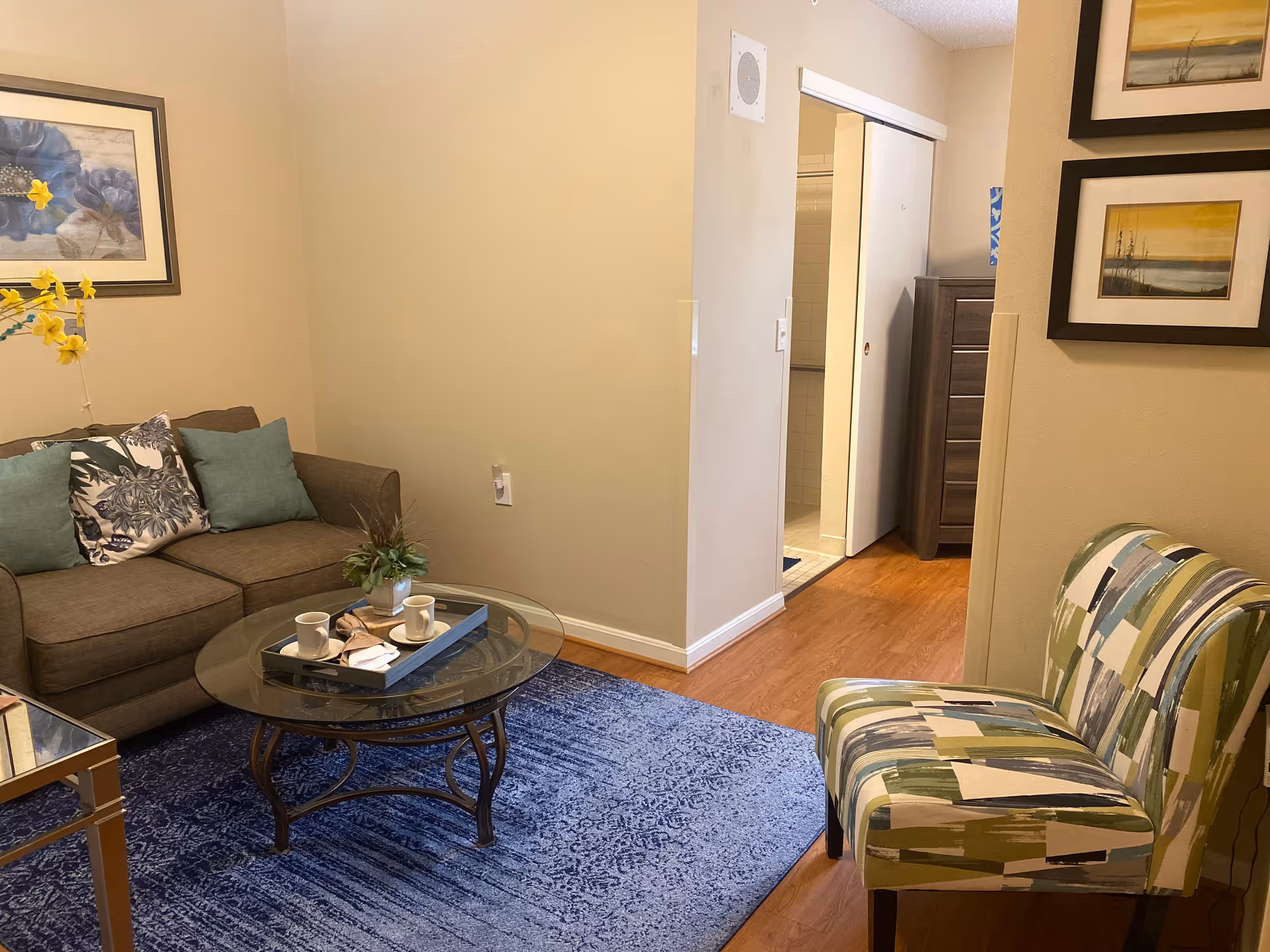 A cozy living room area with a brown sofa adorned with green and floral cushions, a glass coffee table with two cups and a small plant on a tray, a patterned green and blue chair, blue area rug, and framed artwork on beige walls. A doorway leads to a bathroom with a sliding door and a wooden dresser is visible in the hallway.