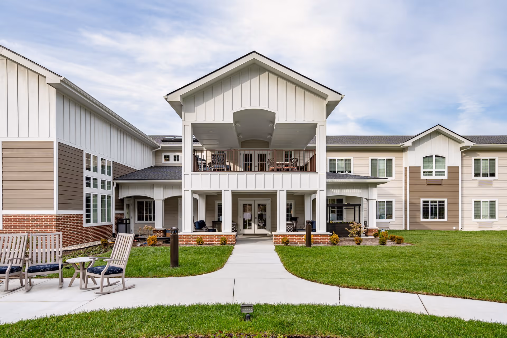 Exterior view of The Vero at Chesapeake Senior Living community building with a covered porch and balcony. The building features white and beige siding with brick accents, large windows, and a well-maintained lawn with outdoor seating including chairs and a small table.