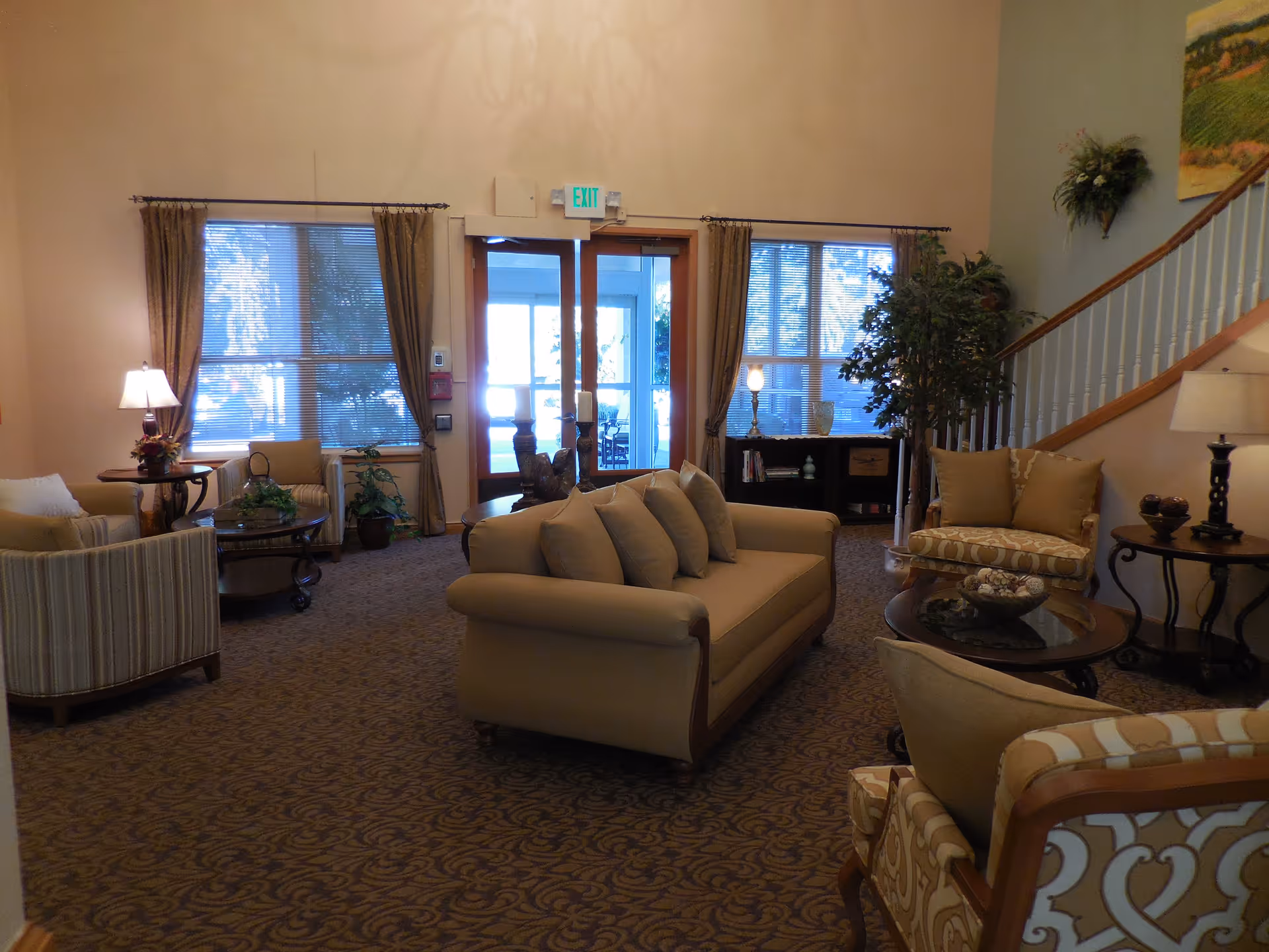 A cozy living room area with beige and patterned armchairs, a beige sofa with multiple cushions, round wooden coffee tables, lamps, potted plants, and large windows with curtains. There is a staircase on the right side and a glass door with an exit sign above it in the background.
