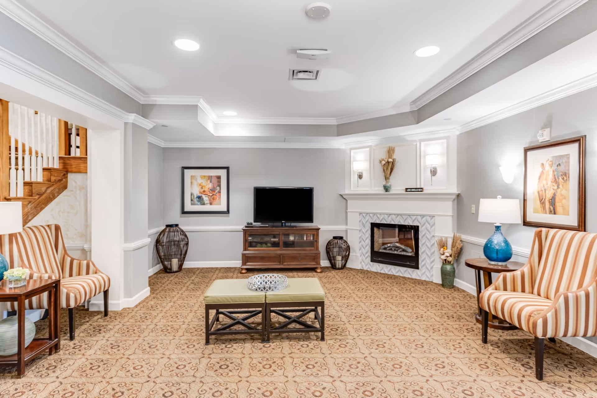A cozy living room with beige patterned carpet, two striped armchairs on either side, a small green ottoman in the center with a decorative tray, a wooden TV stand with a flat-screen TV, and a white fireplace with decorative vases and wall sconces. There are framed paintings on the walls and a staircase visible to the left.