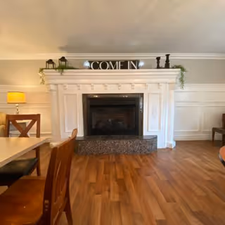 A cozy interior room with a white decorative fireplace centered on the wall. On top of the fireplace mantel, there is a sign that says 'COME IN' along with some small decorative items and greenery. The room has wooden flooring and wooden chairs and tables are partially visible on the left and right sides. A lit table lamp is visible on the left side of the image.
