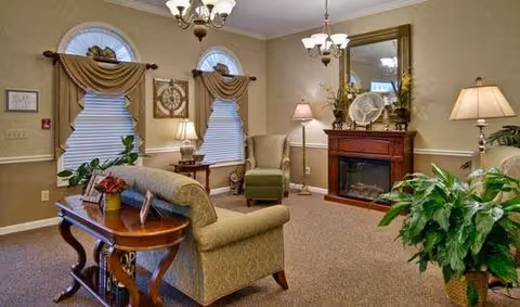 A cozy living room with beige walls and carpet, featuring a green upholstered sofa and armchair, a wooden coffee table with decorative items, a fireplace with a large mirror above it, two arched windows with draped curtains, and several lamps providing warm lighting. There are also green plants adding a touch of nature to the room.