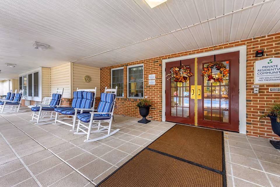 Covered entrance area of a residential community with brick walls and double glass doors decorated with autumn wreaths. Several white rocking chairs with blue cushions are lined up along the wall under the covered porch. A brown doormat is placed in front of the doors, and there are potted plants on either side of the entrance. A sign on the wall indicates it is a private residential community with no soliciting or trespassing.