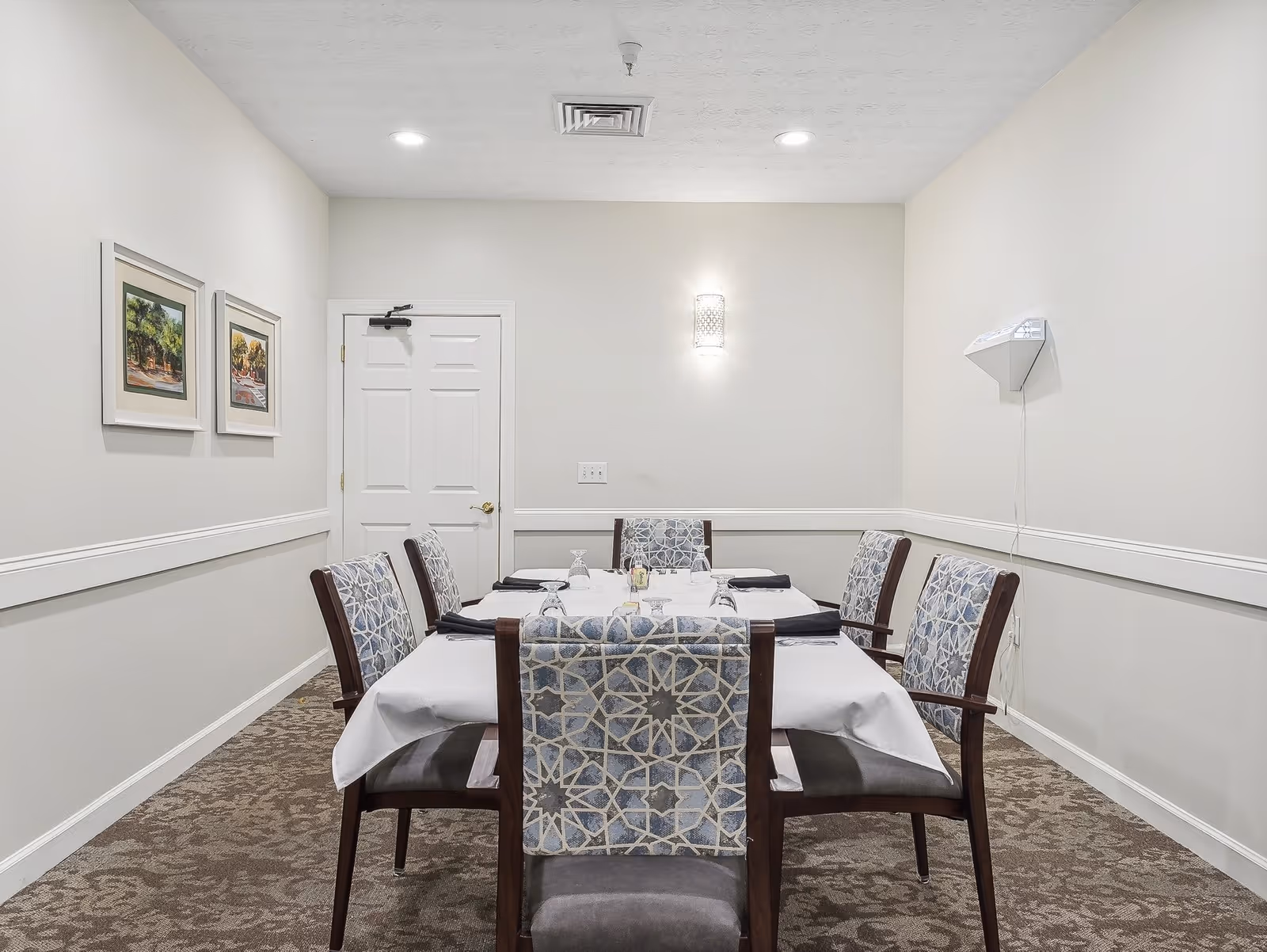 A small dining room with a rectangular table covered with a white tablecloth and six chairs with patterned fabric backs. The room has beige walls, two framed paintings on the left wall, a closed white door at the back, and a wall-mounted light fixture above the table. The floor is carpeted with a patterned design.