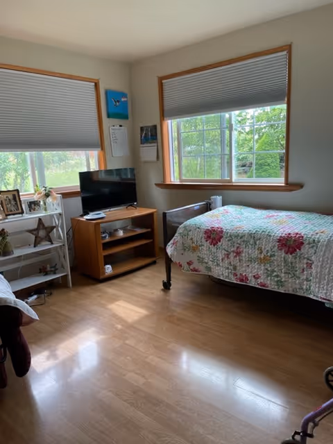 A bright bedroom with wooden flooring, a bed covered with a floral quilt, a small wooden TV stand with a flat-screen TV, and two windows with blinds partially drawn. There is a white shelving unit with picture frames and decorative items near one window.