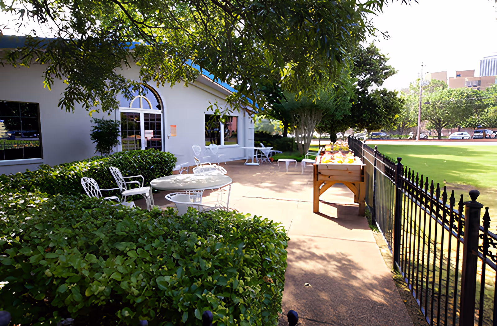 Outdoor patio area at AHC Savannah with white metal tables and chairs surrounded by green bushes and trees. A building with large windows and a door is visible on the left side. A black metal fence runs along the right side, bordering a green lawn area with parked cars and buildings in the background.