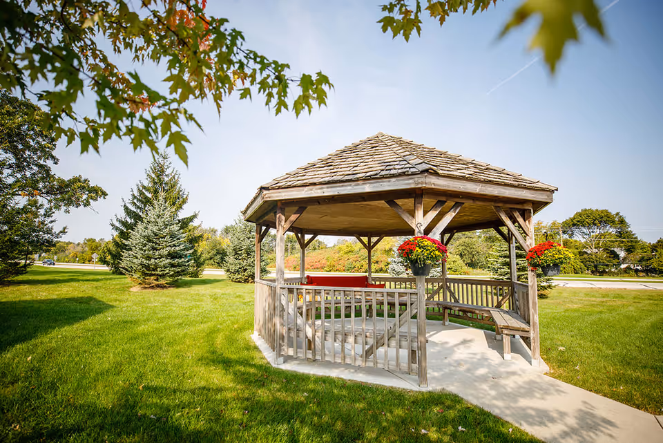 A wooden gazebo with a shingled roof situated on a green lawn surrounded by trees and shrubs under a clear blue sky. The gazebo has benches inside and hanging flower pots with red and yellow flowers.