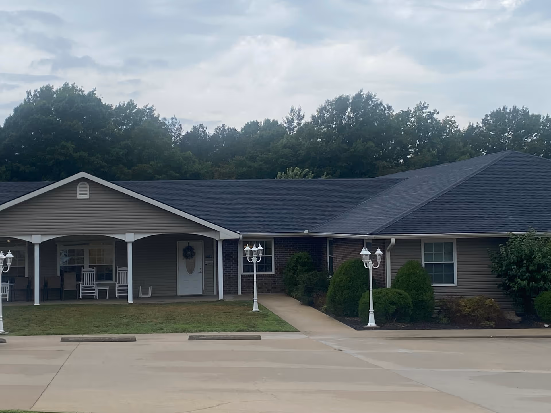 Exterior view of Ambrose Park Residential Care, showing a single-story building with a covered porch, white rocking chairs, two white lamp posts, and surrounding greenery with trees in the background under a cloudy sky.