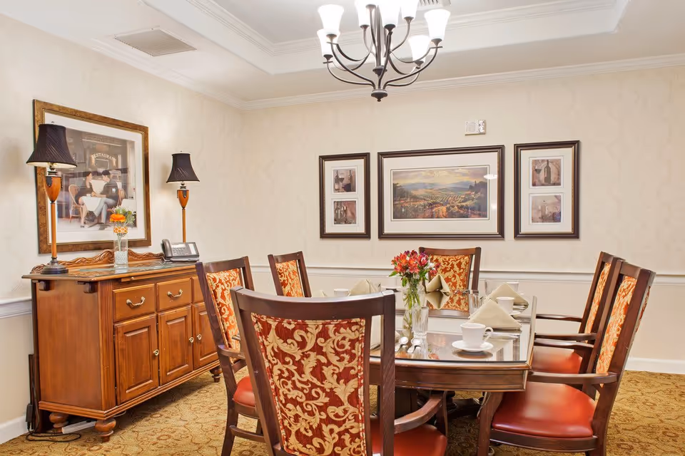 A formal dining room with a wooden dining table surrounded by six chairs with red and gold patterned upholstery. The table is set with white cups, glasses, and beige napkins. A vase with red flowers is placed in the center. On the left side, there is a wooden sideboard with two lamps and a framed picture above it. The walls are decorated with three framed pictures, and a chandelier hangs from the ceiling.
