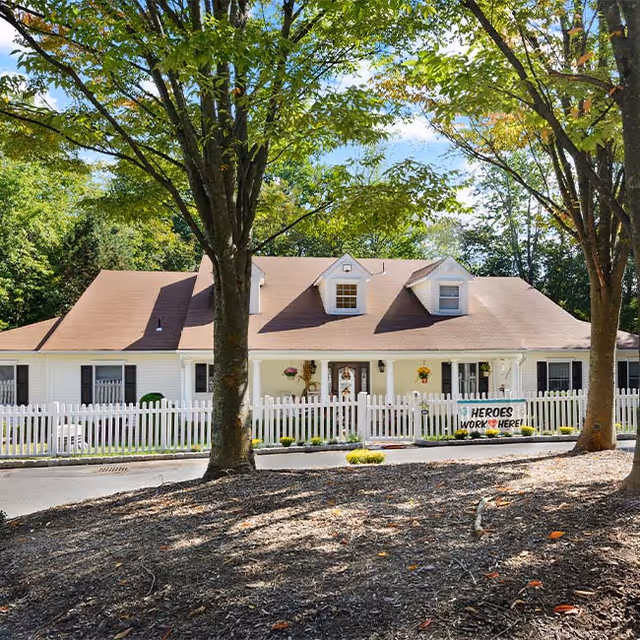 A single-story white building with a brown roof surrounded by a white picket fence. There are two large trees in the foreground and a sign on the fence that reads 'HEROES WORK HERE'. The building is set against a backdrop of green trees under a blue sky with some clouds.
