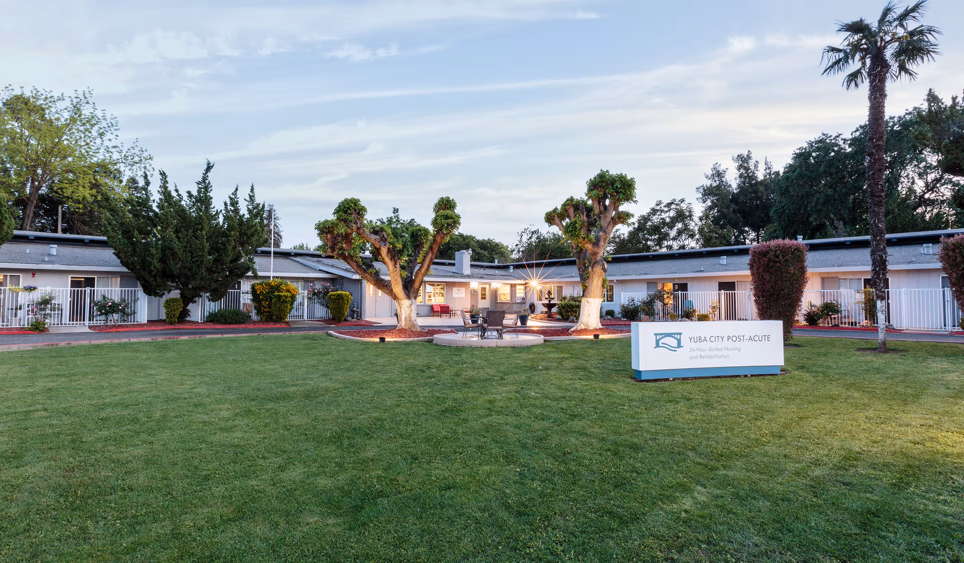 Exterior front lawn and courtyard of Yuba City Post-Acute with trimmed trees, a central seating area, and a facility sign.