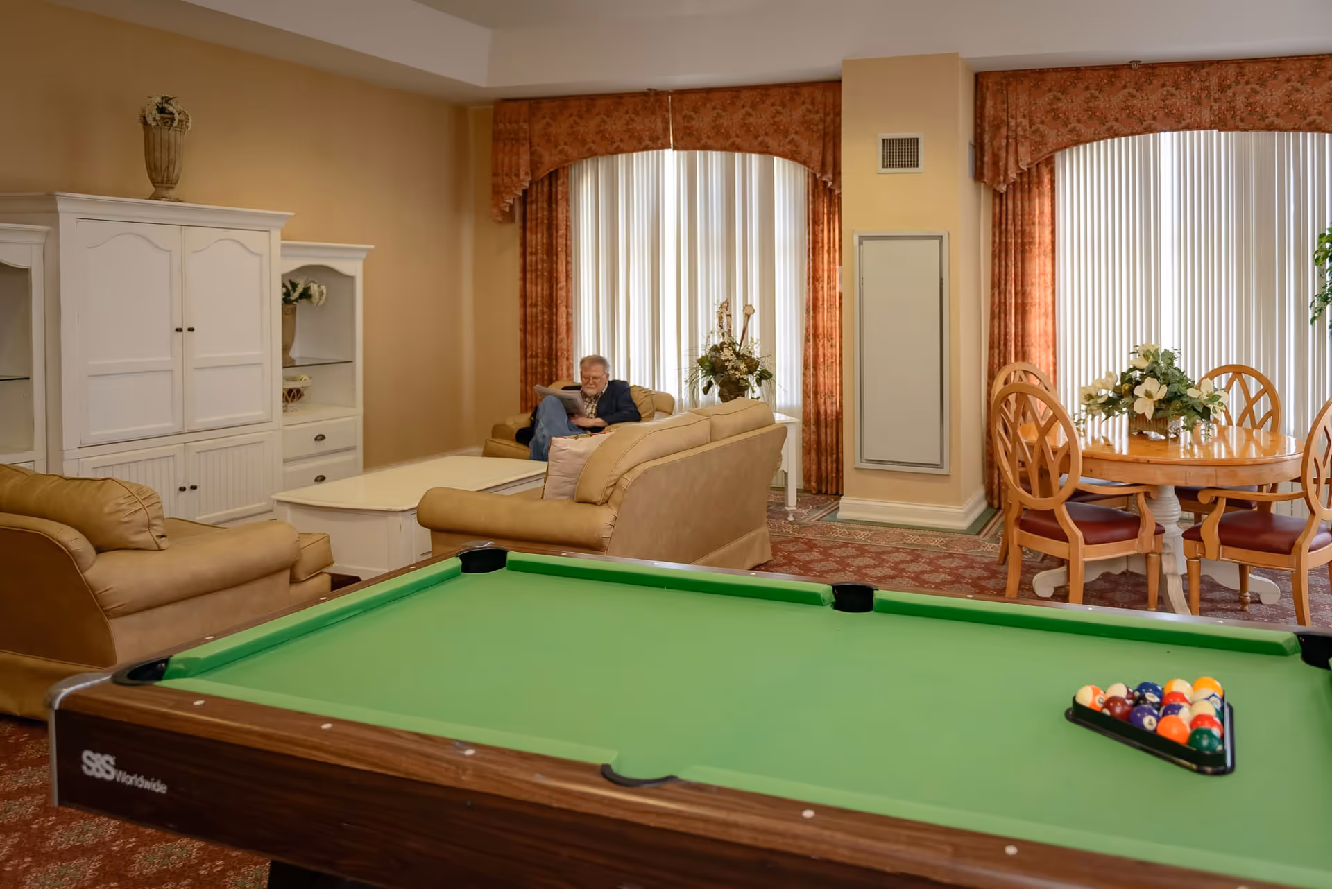 Interior common room with a green pool table in the foreground, sofas and a dining table by tall windows, and a person seated on a couch.