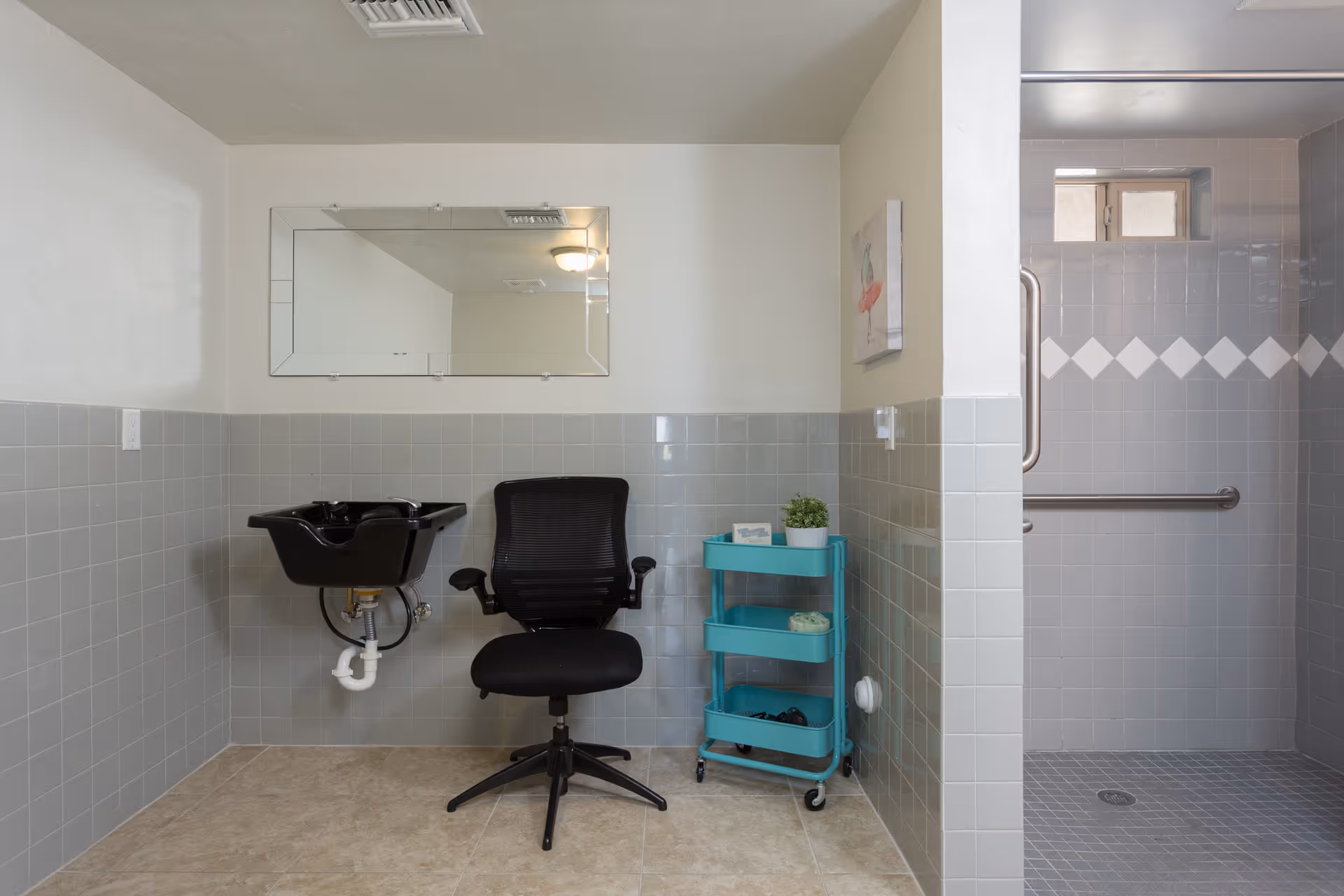 A small hair washing station in a tiled room with a black salon chair, a black sink, a large rectangular mirror on the wall, and a turquoise rolling cart with various items. To the right is a tiled walk-in shower with grab bars and a small window.