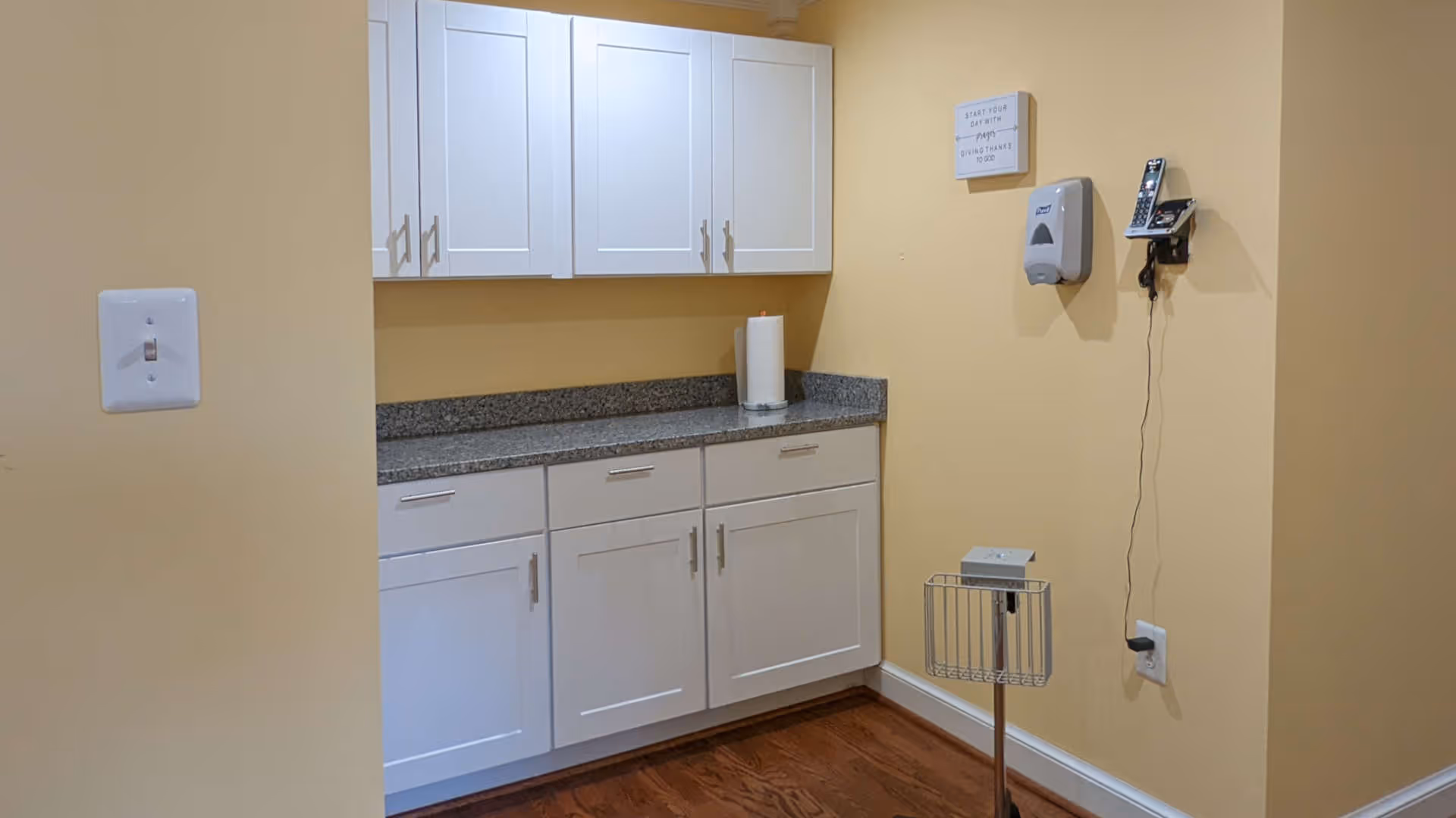 A small kitchenette area with white cabinets, a granite countertop, and yellow walls. There is a paper towel roll on the counter, a wall-mounted phone, a hand sanitizer dispenser, and a small metal stand with a device on it. The floor is wooden.