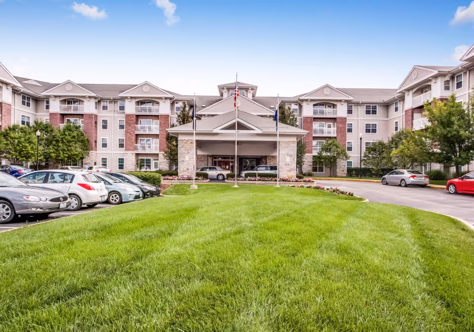 Front exterior view of a multi-story senior living facility with a covered entrance, three flagpoles, well-maintained green lawn, parked cars, and trees on either side.