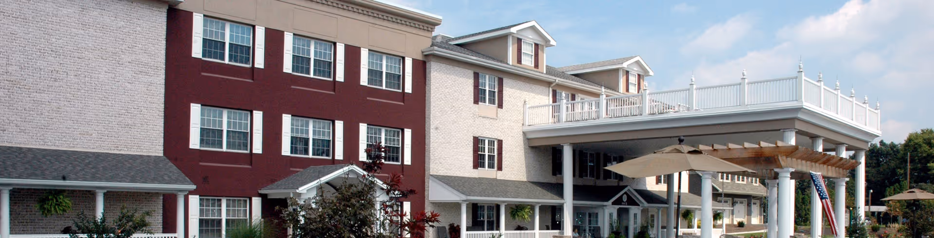 Exterior view of a multi-story senior living facility building with white and maroon brick walls, multiple windows, a covered entrance with white columns, and a pergola. There are some plants and trees in front of the building under a partly cloudy sky.