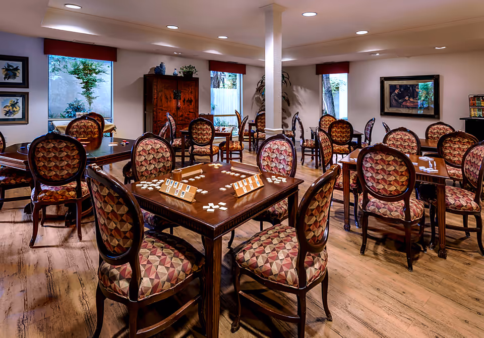 A well-lit room with multiple wooden tables and chairs featuring patterned upholstery. The tables have game tiles and racks set up, suggesting a game or activity room. The room has large windows with red valances, framed artwork on the walls, and wooden flooring.