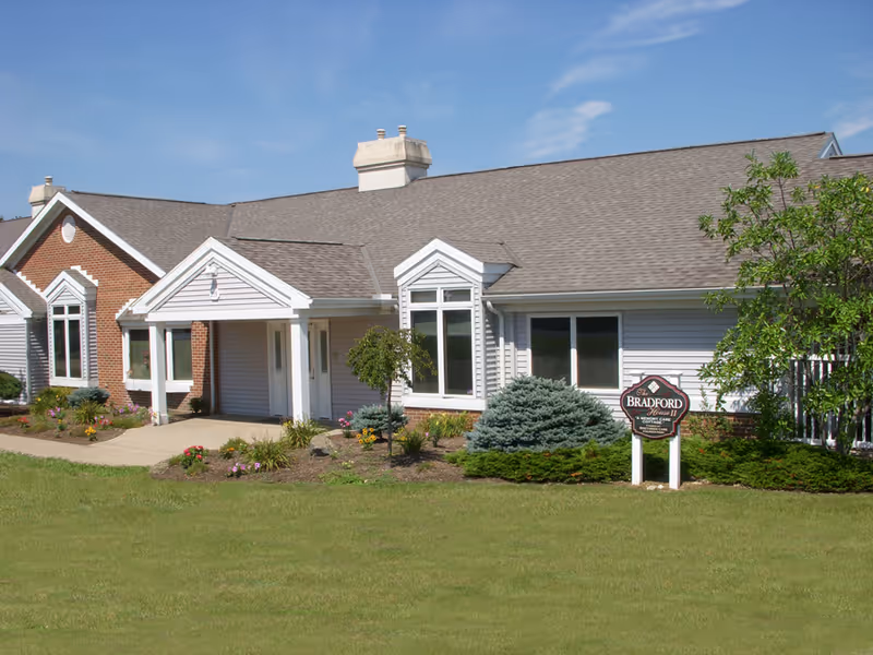 Exterior view of a single-story building with a gabled roof, white siding, and brick accents. The building has large windows and a covered entrance with white columns. There is a well-maintained lawn and landscaped garden with shrubs and flowers in front. A sign near the entrance reads 'Bradford House II'.