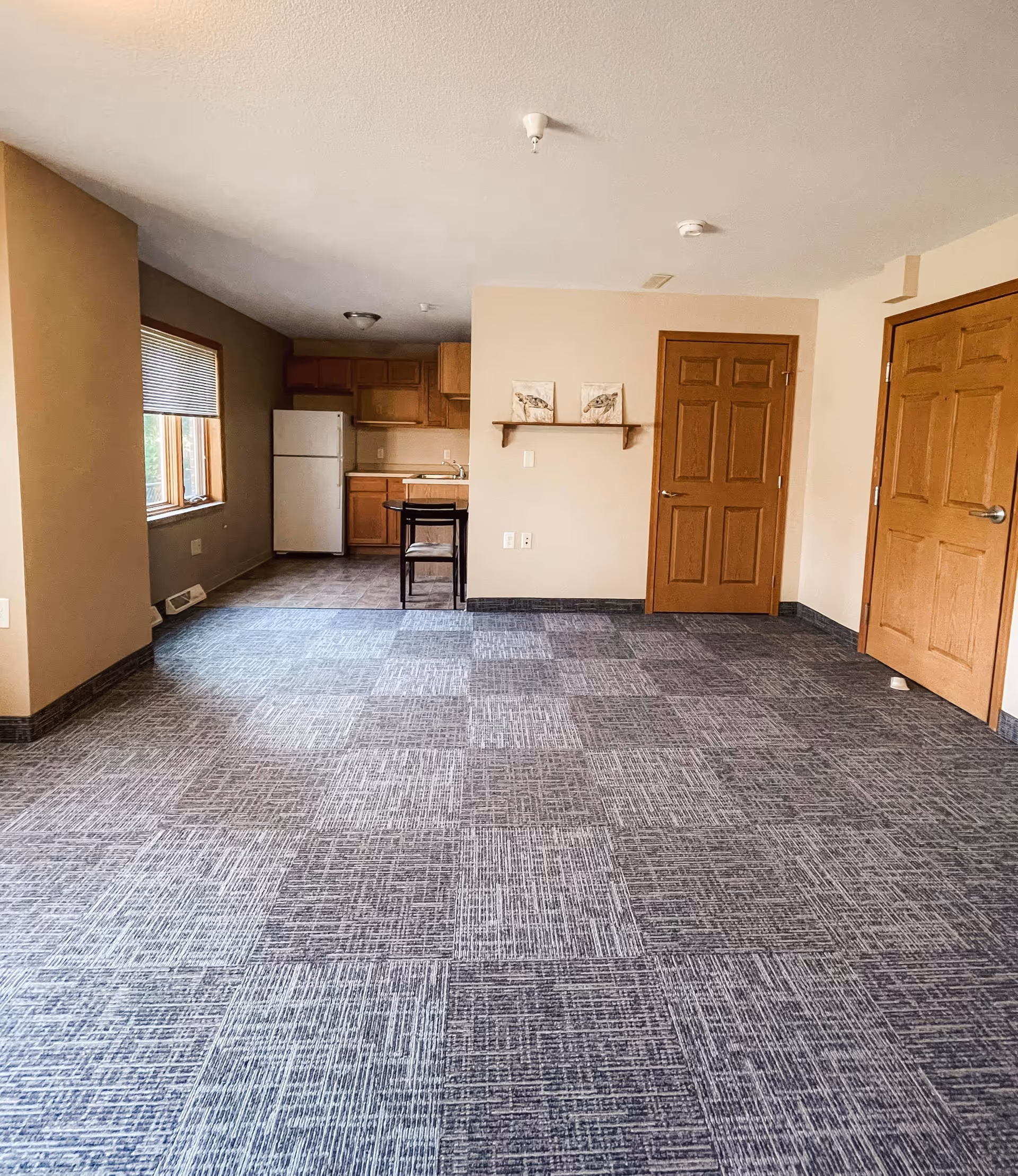 An empty room with patterned carpet flooring, beige walls, and two wooden doors on the right side. At the back of the room is a small kitchen area with wooden cabinets, a white refrigerator, a sink, and a small table with two chairs. A window with blinds is on the left wall, allowing natural light into the space.