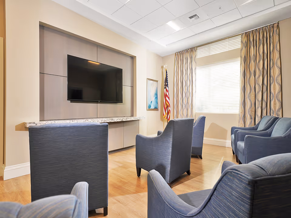 A bright communal sitting room with blue upholstered armchairs arranged facing a wall-mounted flat-screen TV, a large window with patterned curtains, and an American flag.
