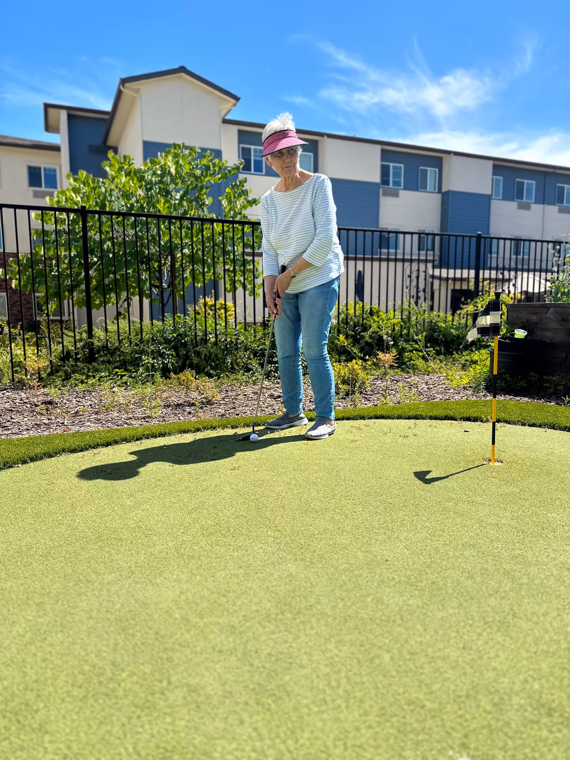 An elderly woman wearing a pink visor, striped long-sleeve shirt, and jeans is playing mini golf on a putting green outdoors. Behind her is a black metal fence, green bushes, and a multi-story building under a clear blue sky.