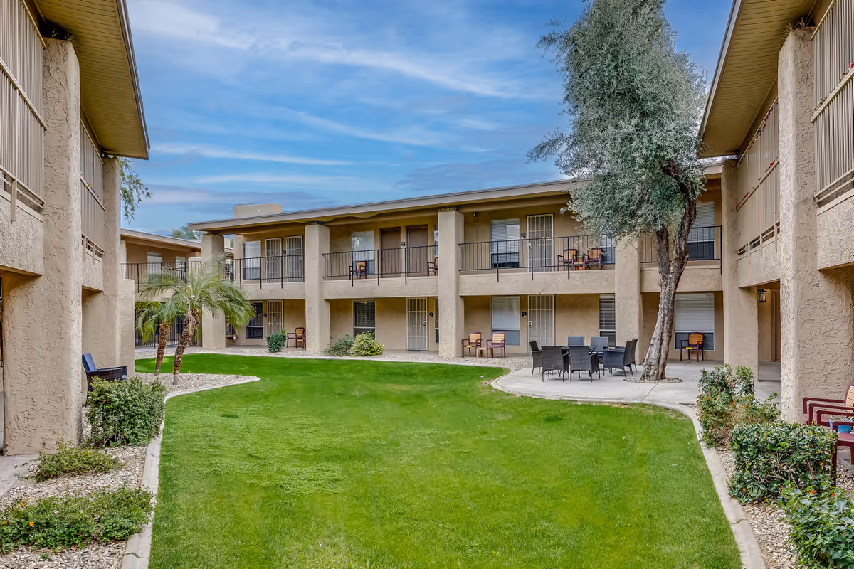 Outdoor courtyard area of Scottsdale Village Square featuring a well-maintained green lawn, a few palm trees, and a large tree near a circular concrete patio with a table and chairs. The courtyard is surrounded by a two-story beige building with balconies and patio chairs.