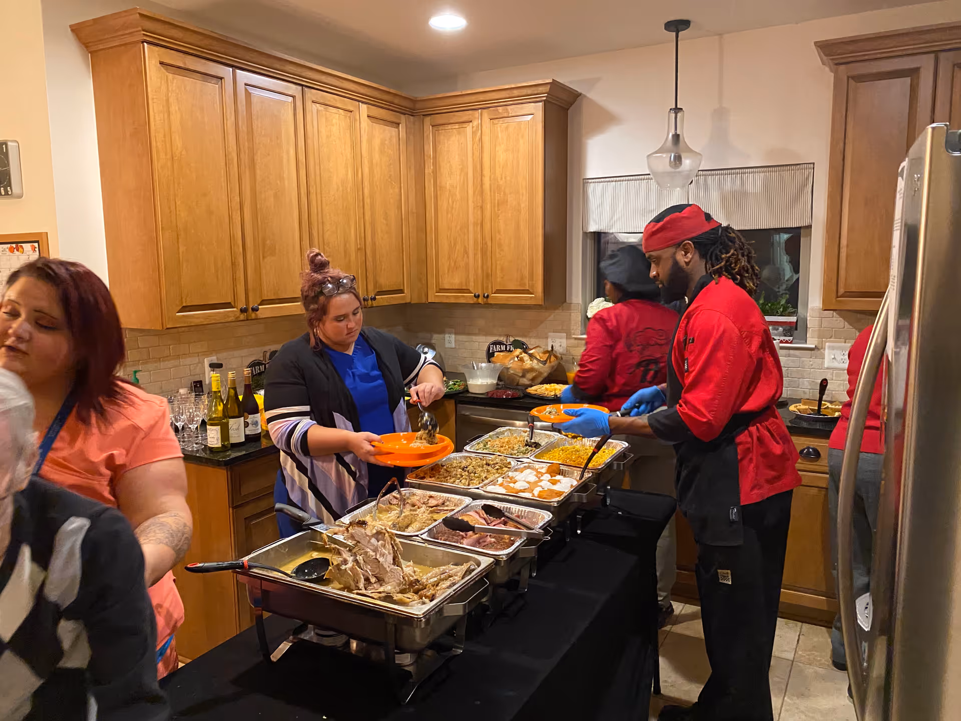 People serving themselves food from a buffet set up in a kitchen with wooden cabinets. Various dishes are laid out in metal trays on a black tablecloth-covered table. A man in a red chef uniform is serving food to a woman holding an orange plate.