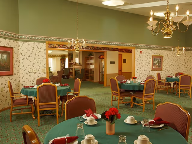 Dining room with round tables covered in green tablecloths, place settings, red napkins and floral centerpieces under chandeliers.