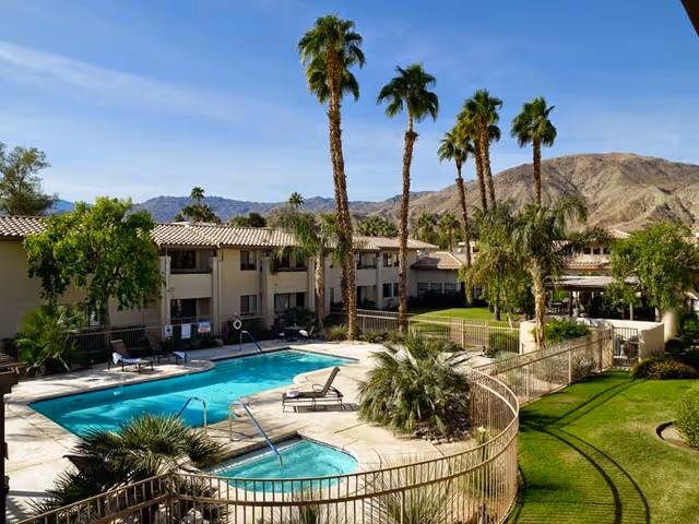 Outdoor view of a senior living facility with a swimming pool and a hot tub surrounded by a safety fence. There are lounge chairs around the pool, palm trees, green lawns, and a two-story building in the background with mountains visible further behind under a clear blue sky.
