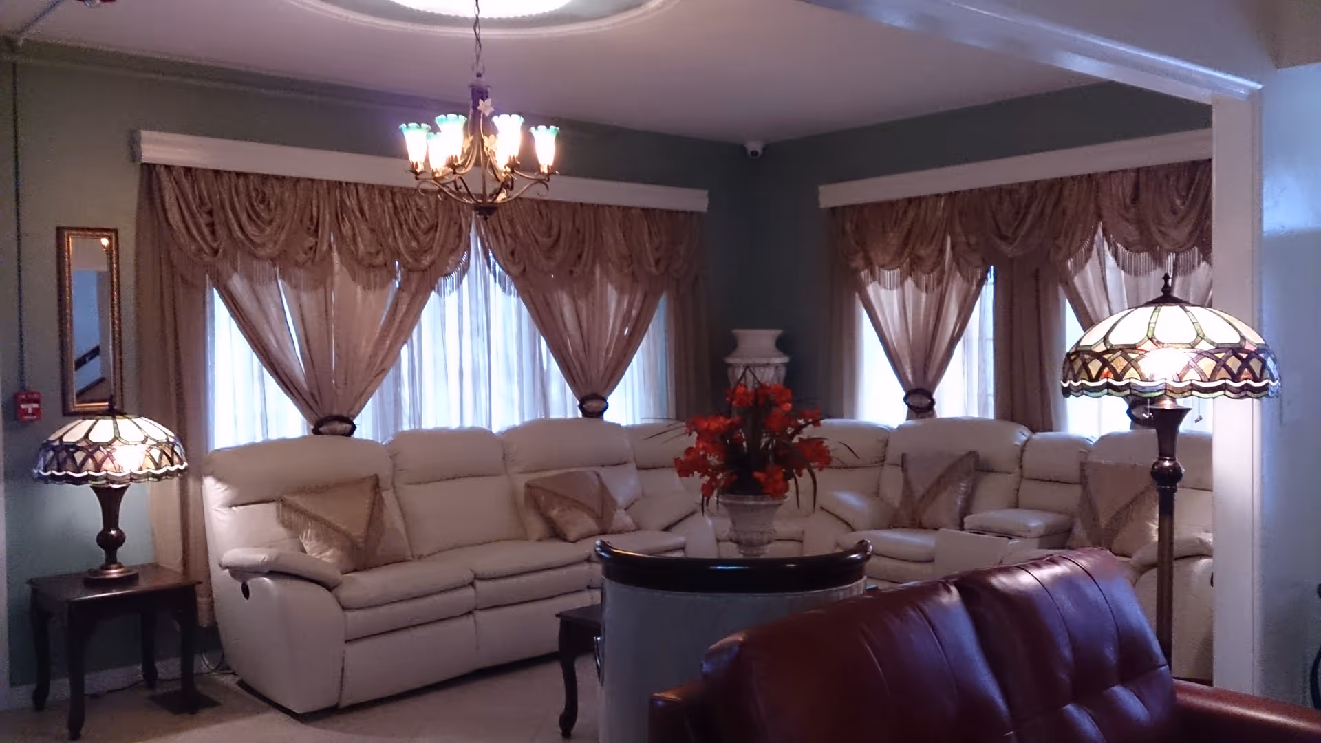 A cozy living room with a large beige sectional sofa adorned with matching pillows, a dark wooden side table with a decorative lamp, and a chandelier hanging from the ceiling. The room has green walls with large windows covered by sheer curtains and ornate valances. A red flower arrangement sits on a round table in the center, and a brown leather chair is partially visible in the foreground.