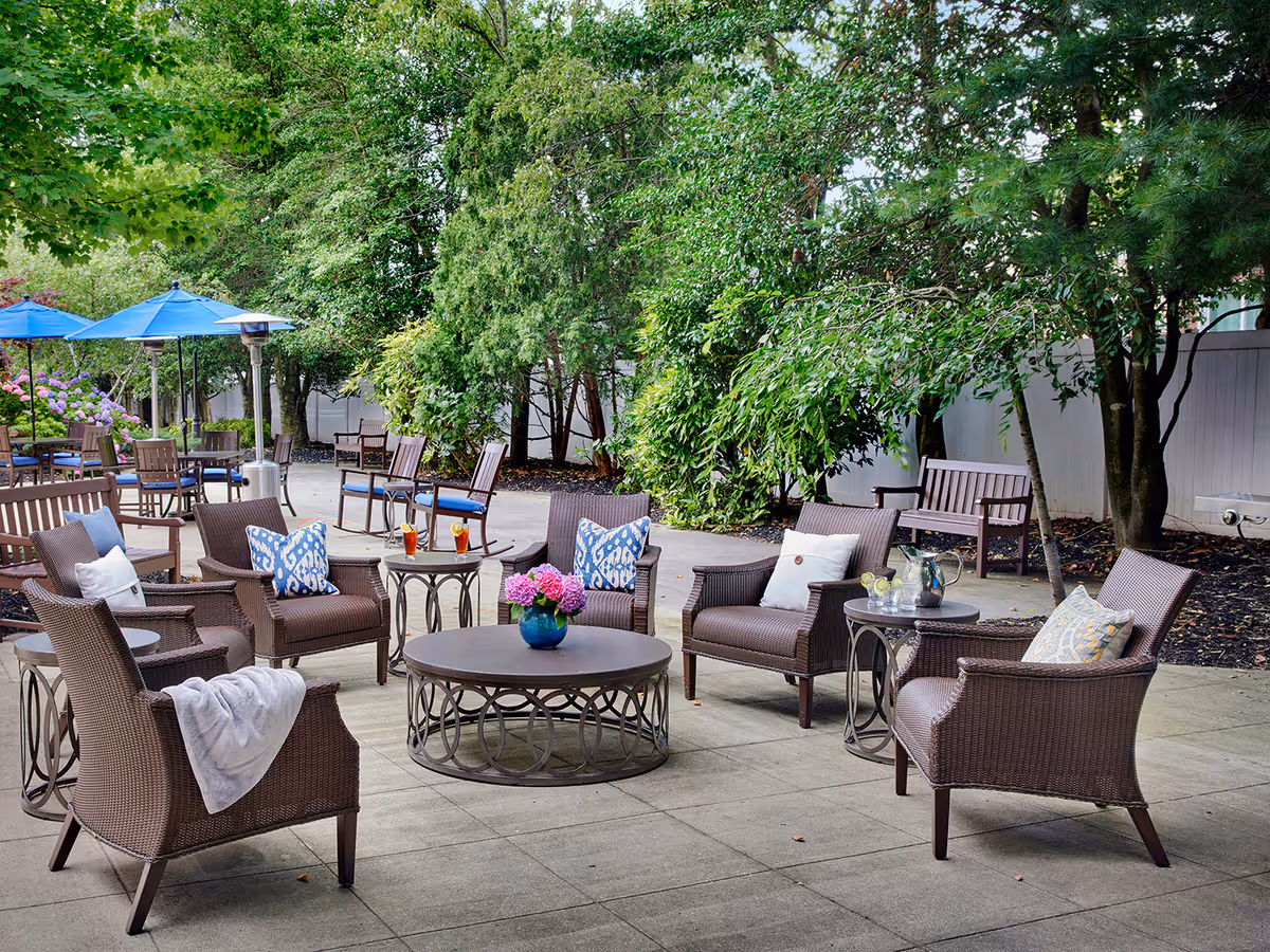 Outdoor patio area with brown wicker chairs and tables arranged in a circle, decorated with cushions and a vase of flowers on the central table. The patio is surrounded by green trees and plants, with additional seating and blue umbrellas in the background.