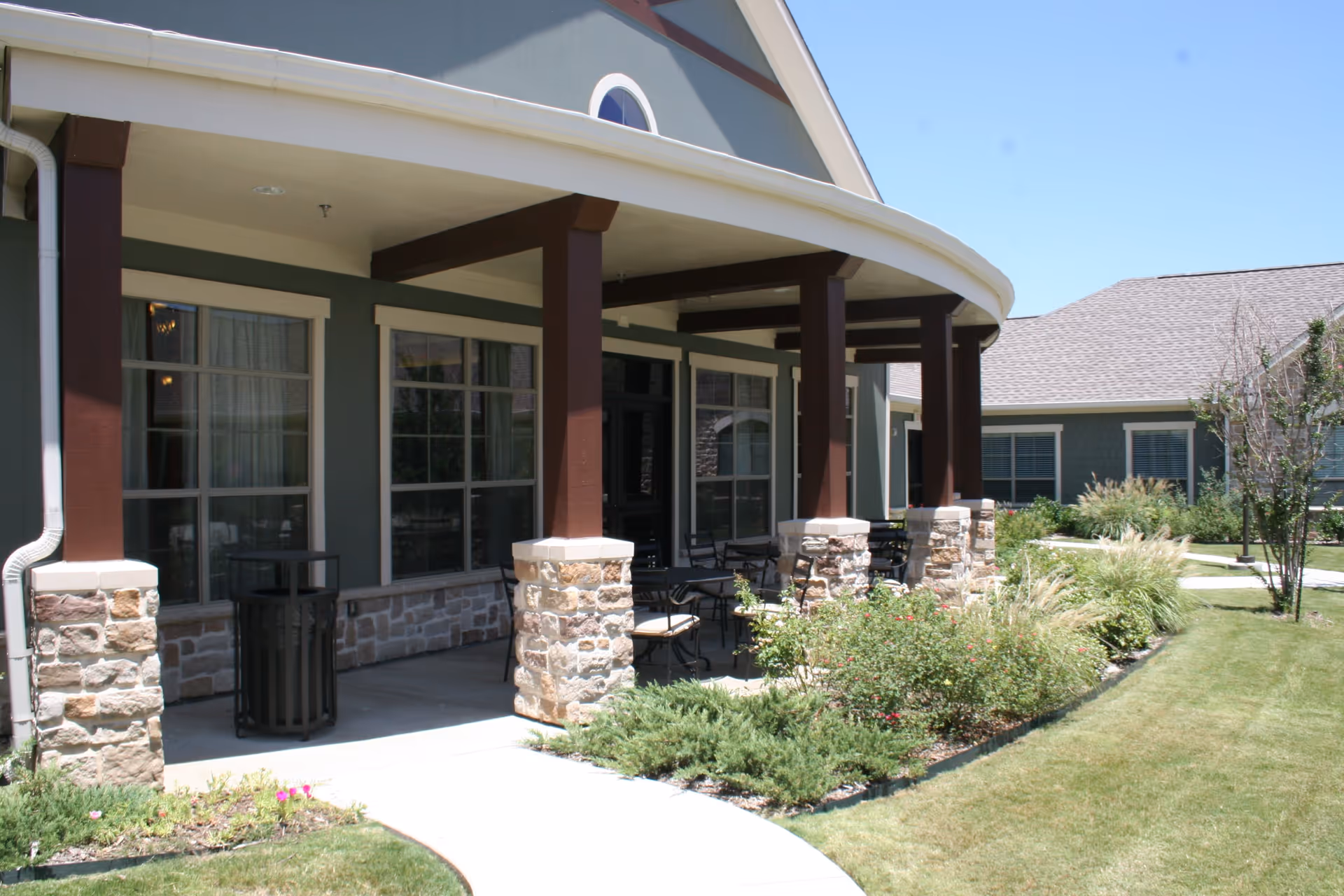 Outdoor covered patio area at Heritage Place Assisted Living & Memory Care with stone pillars, brown wooden beams, several chairs and tables, and landscaped garden beds with shrubs and flowers along a curved concrete walkway.