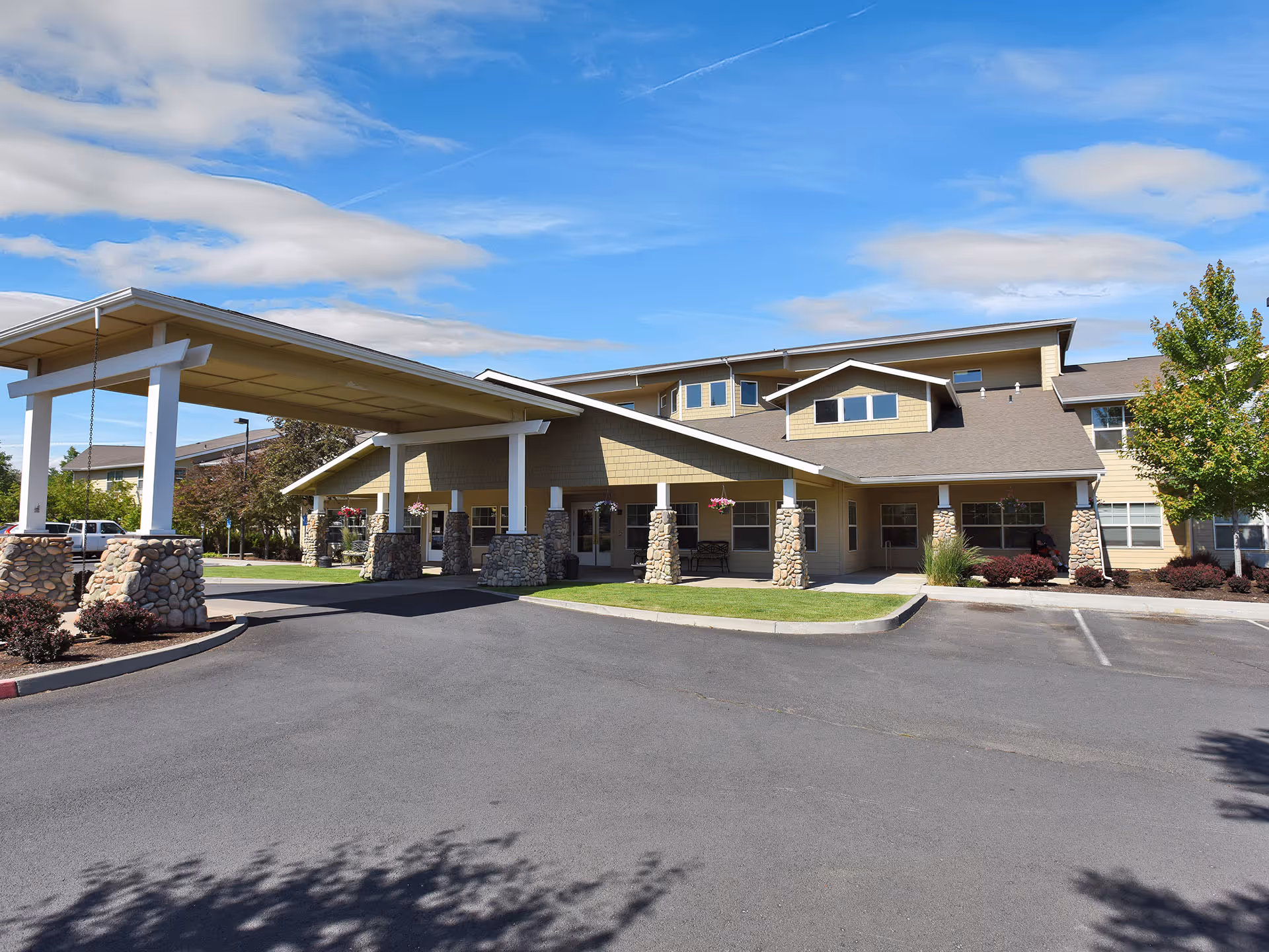 Exterior front view of Prestige Senior Living High Desert building with a covered entrance supported by stone pillars, surrounded by a parking area and landscaped greenery under a partly cloudy blue sky.