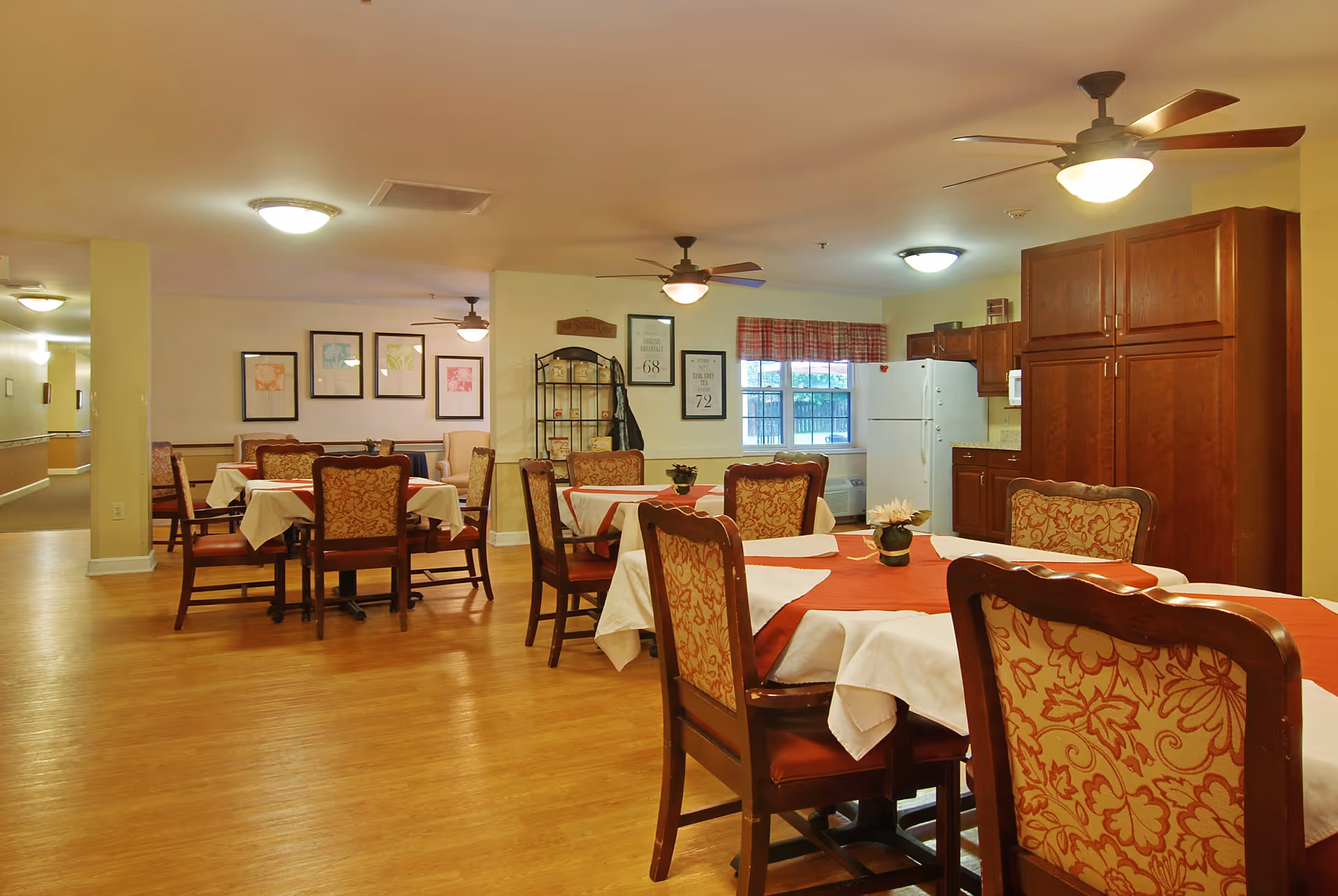 A dining area in a senior living facility with several tables covered with white and red tablecloths. Each table is surrounded by wooden chairs with patterned upholstery. The room has wooden flooring, ceiling fans with lights, framed artwork on the walls, and a kitchen area with wooden cabinets and a white refrigerator in the background.