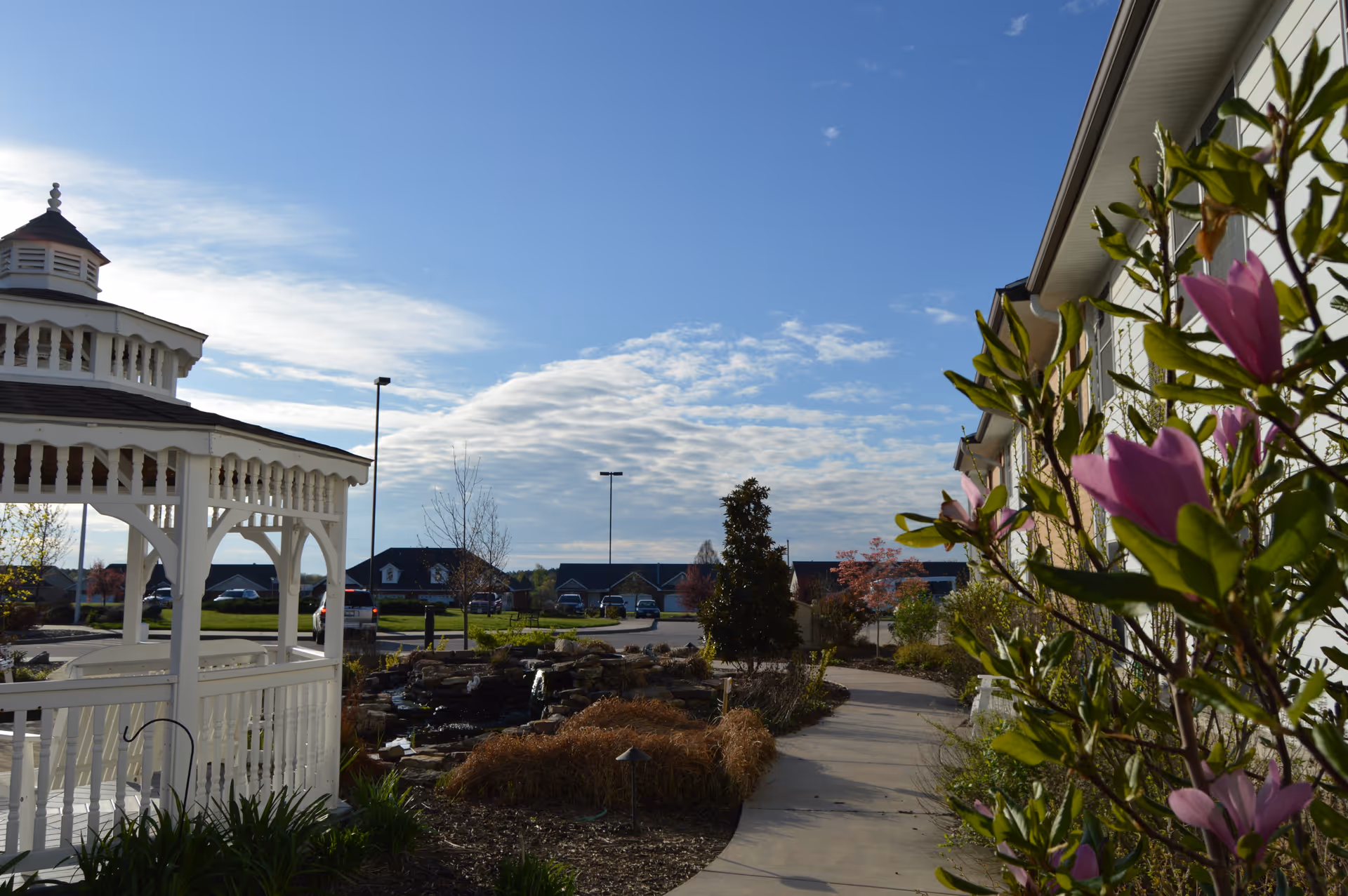 Outdoor view of Magnolia Terrace Senior Living Apartments featuring a white gazebo on the left, a landscaped garden with a small pond and plants, a paved walkway, and a building with flowering bushes on the right under a partly cloudy blue sky.