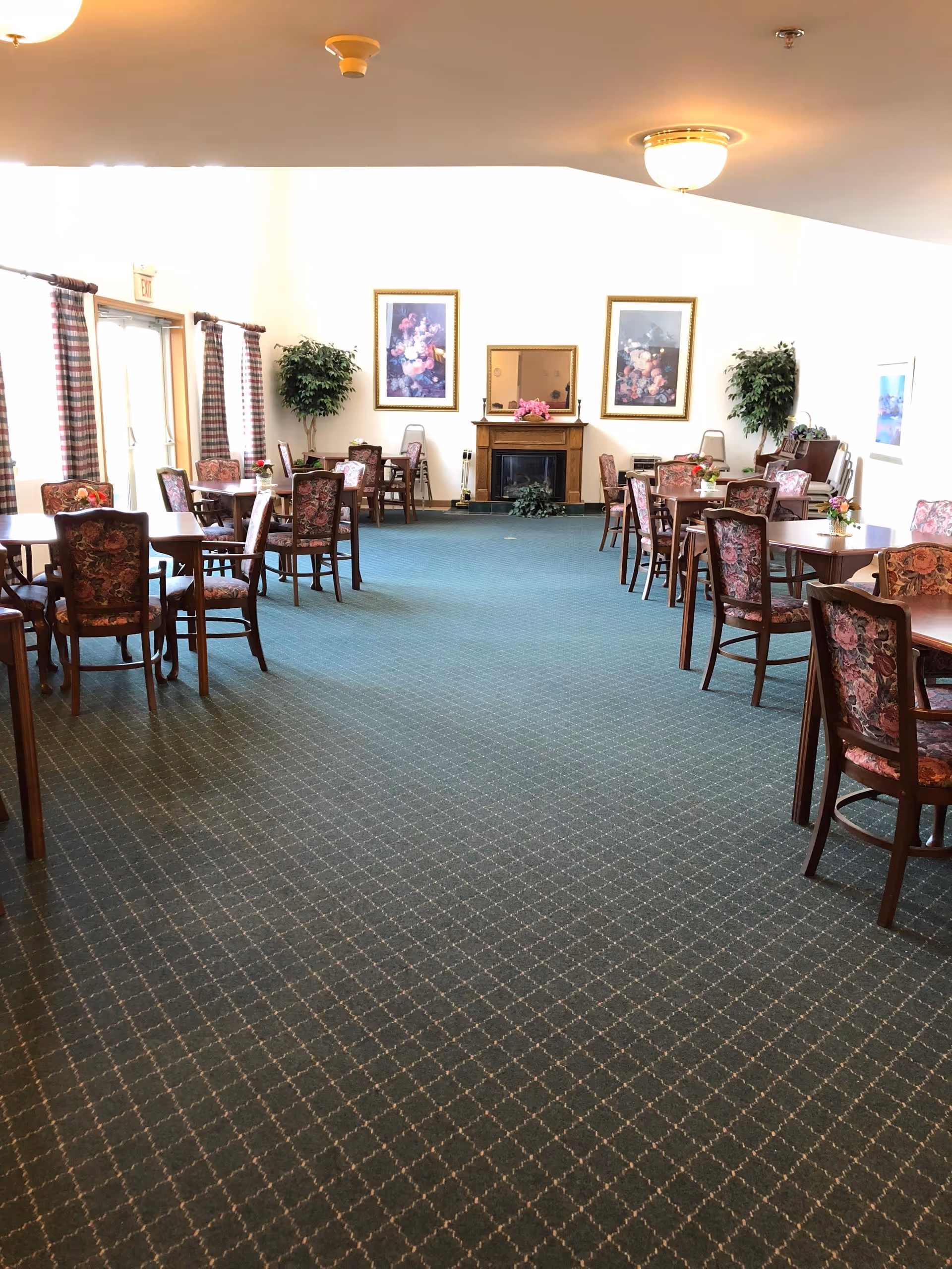 A spacious dining room with multiple wooden tables and chairs upholstered in floral fabric. The room has a green carpet with a diamond pattern, two large framed floral paintings on the far wall, a fireplace with a mirror above it, and two large potted plants on either side. There are large windows with plaid curtains on the left side, allowing natural light to enter the room.