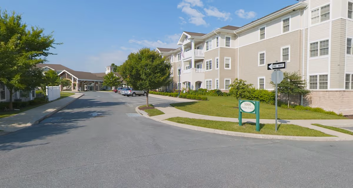Exterior view of Carroll Lutheran Village showing a paved driveway with parked cars, green lawns, trees, and multi-story beige and white buildings under a blue sky with some clouds.