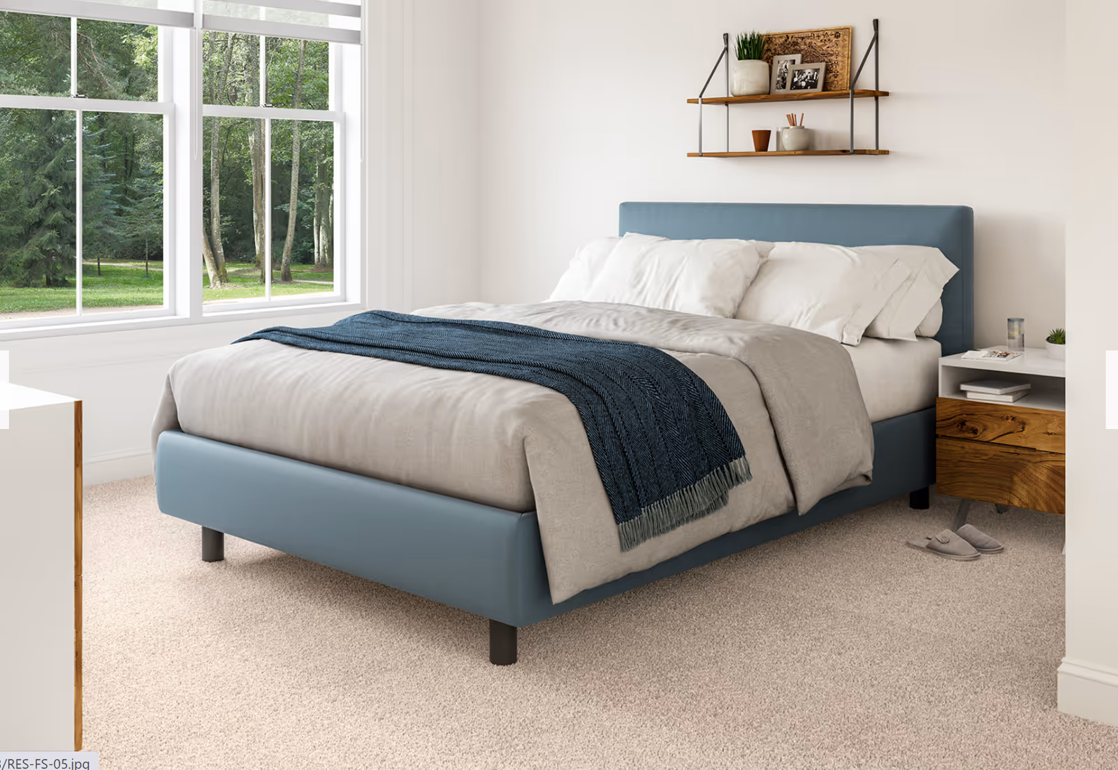 A cozy bedroom with a large window showing a green forest outside. The room features a blue upholstered bed with beige bedding and a dark blue throw blanket. There is a wooden nightstand with books, a glass of water, and a small plant. Above the bed, a wall-mounted shelf holds decorative items and framed photos. The floor is carpeted, and a pair of gray slippers are placed beside the nightstand.