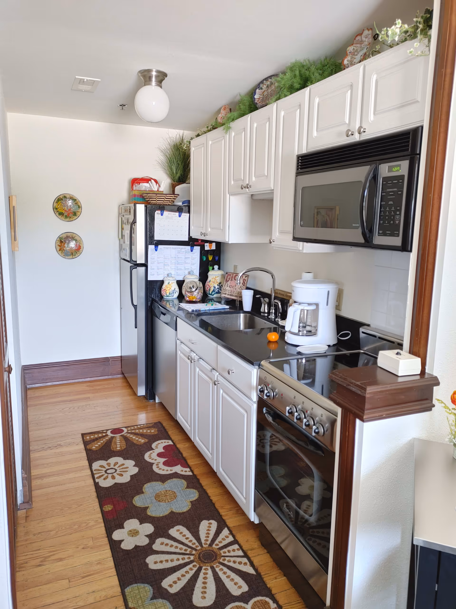 A narrow kitchen with white cabinets, a black countertop, and stainless steel appliances including a refrigerator, dishwasher, stove, and microwave. There is a colorful floral rug on the wooden floor, decorative plates on the wall, and various kitchen items on the counter such as a coffee maker and jars. Greenery and decorative items are placed above the cabinets.