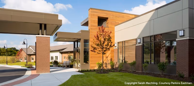 Exterior view of a modern senior living facility with a covered entrance, brick and wood paneling, large windows, a small tree with autumn leaves, and a well-maintained lawn under a partly cloudy sky.