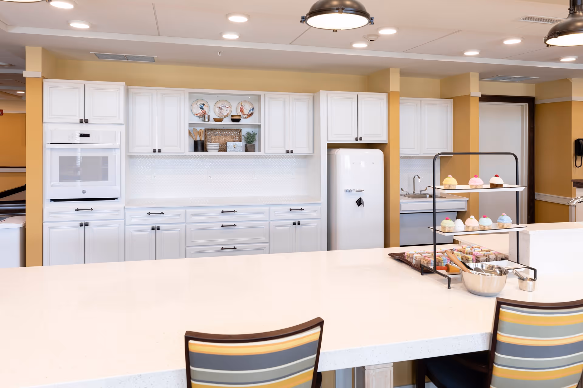 A bright and clean kitchen area with white cabinetry, a white oven, a white retro-style refrigerator, and a white countertop island with two striped chairs. On the countertop, there is a two-tiered stand displaying colorful cupcakes and a tray with various snacks and utensils. The walls are painted a warm yellow color.
