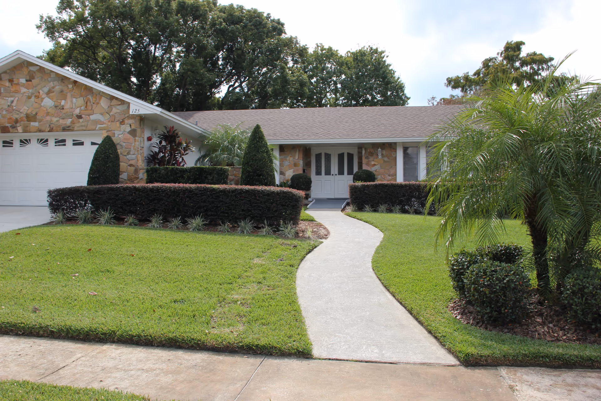 Front exterior view of a single-story house with a stone facade, white double front doors, a garage on the left, a curved concrete walkway leading to the entrance, and well-maintained green lawn and shrubs.