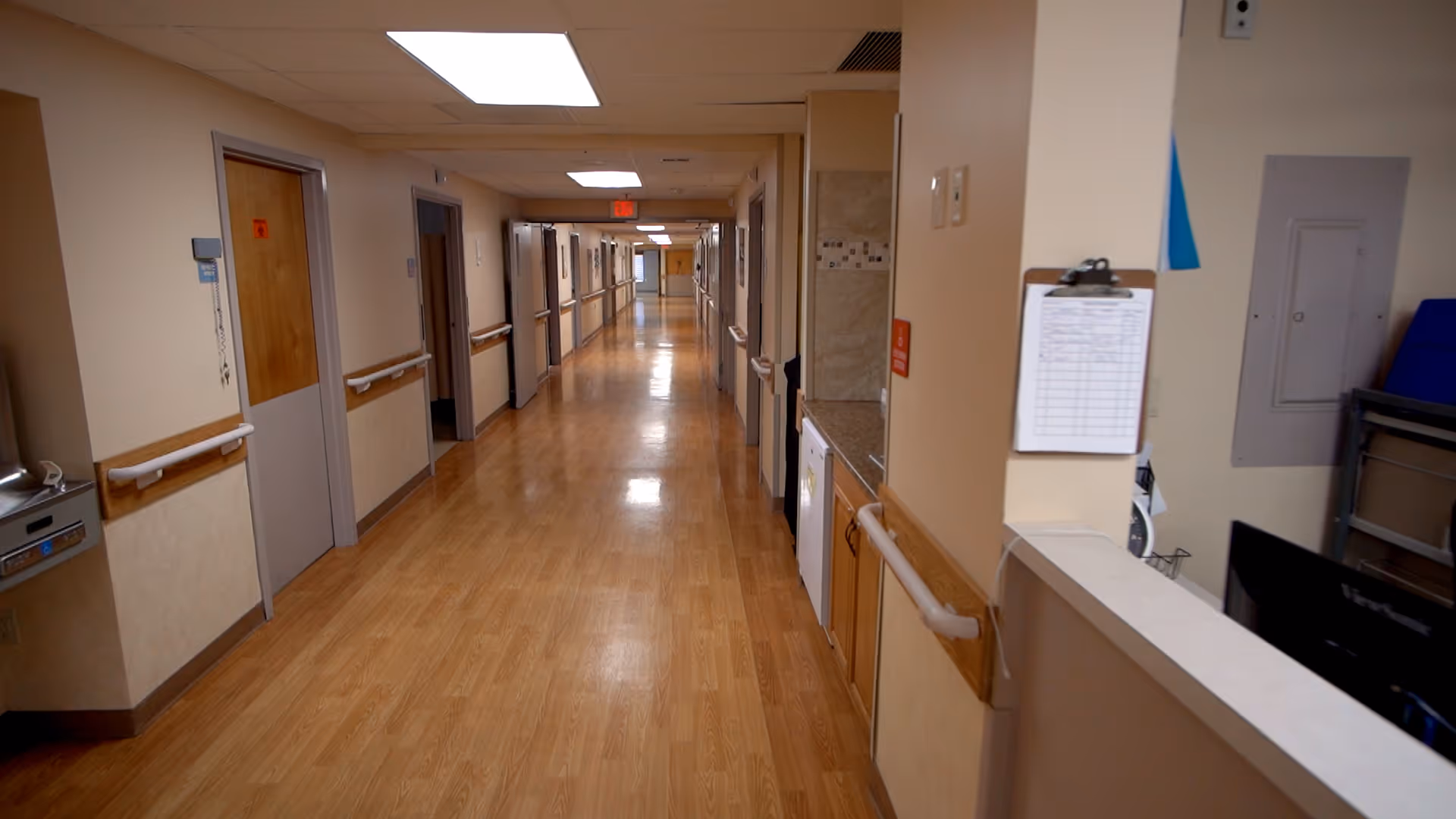 A long, clean hallway in a senior living facility with wooden floors, beige walls, handrails along both sides, multiple closed doors, and bright overhead lighting.