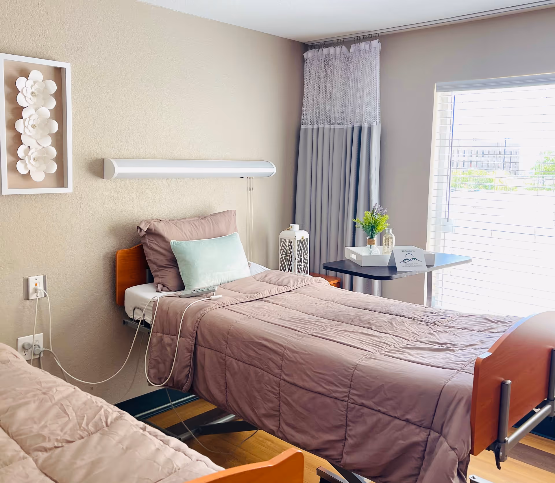 A bright and clean bedroom in a senior living facility with two hospital-style beds. Each bed has mauve bedding and pillows, with one bed featuring a light green accent pillow. A small table near the window holds a white tray with a small plant, a bottle, and a welcome card. The room has beige walls, a window with blinds, and a decorative white flower wall art piece.