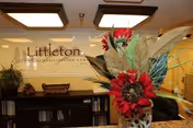 Reception area of Littleton Care and Rehabilitation facility with a decorative flower arrangement in the foreground and the facility's name displayed on the wall behind the reception desk.